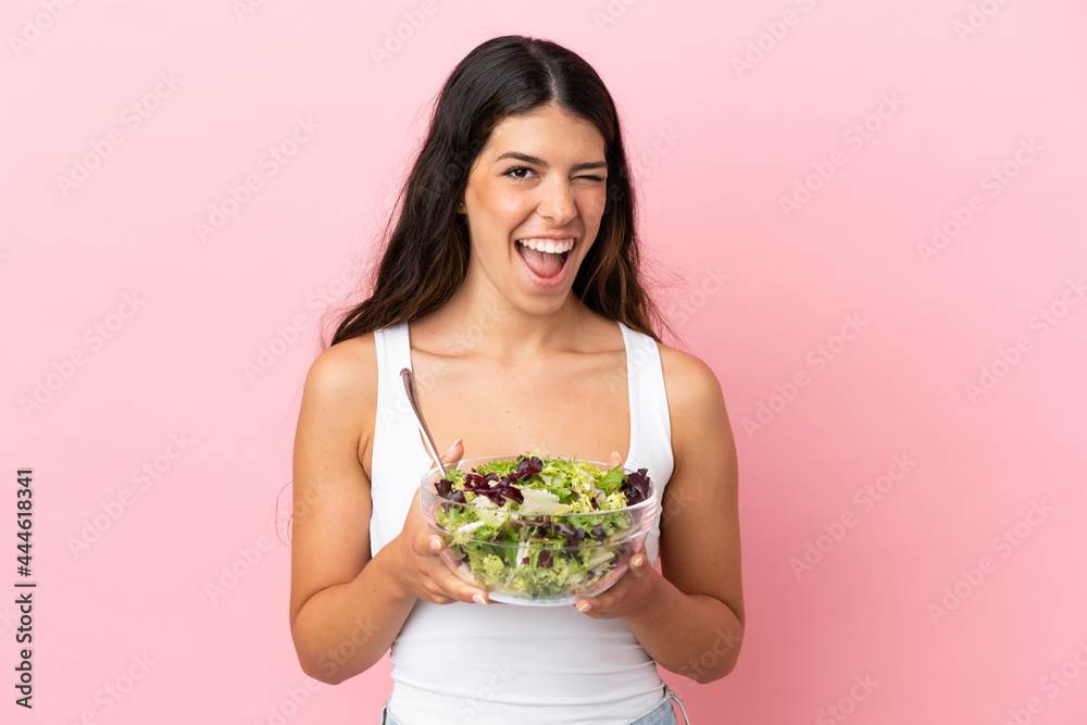 Young caucasian woman isolated on pink background holding a bowl of salad while winking