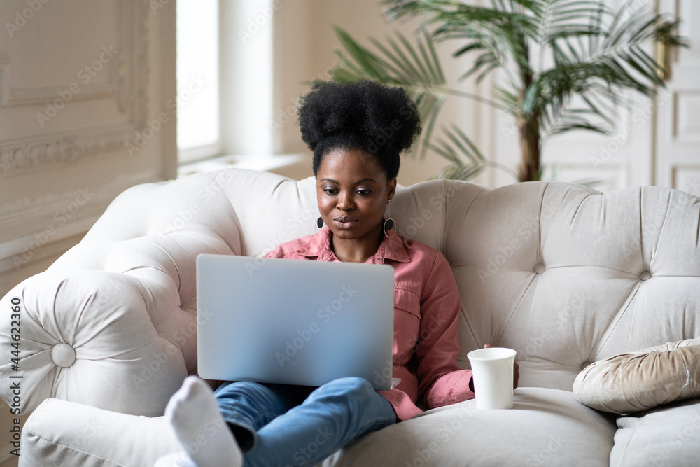 Serious african american woman work on laptop sit on comfortable couch with coffee or tea relaxing after remote freelance occupation from home. Black female read email or browse internet using wifi Serious african american woman work on laptop sit on comfortable couch with coffee or tea relaxing after remote freelance occupation from home. Black female read email or browse internet using wifi
