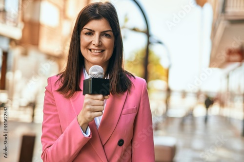 Middle age reporter woman holding microphone doing television speech outdoors