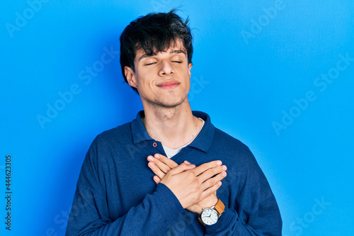 Handsome hipster young man stranding over blue background smiling with hands on chest with closed eyes and grateful gesture on face. health concept.