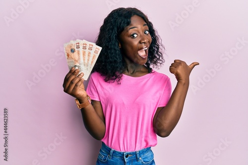 Beautiful african young woman holding 10 united kingdom pounds banknotes pointing thumb up to the side smiling happy with open mouth photo