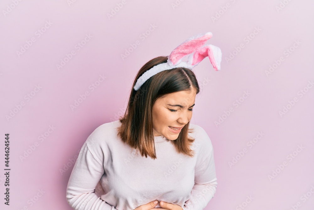 Young beautiful woman wearing cute easter bunny ears with hand on stomach because nausea, painful disease feeling unwell. ache concept. Young beautiful woman wearing cute easter bunny ears with hand on stomach because nausea, painful disease feeling unwell. ache concept.