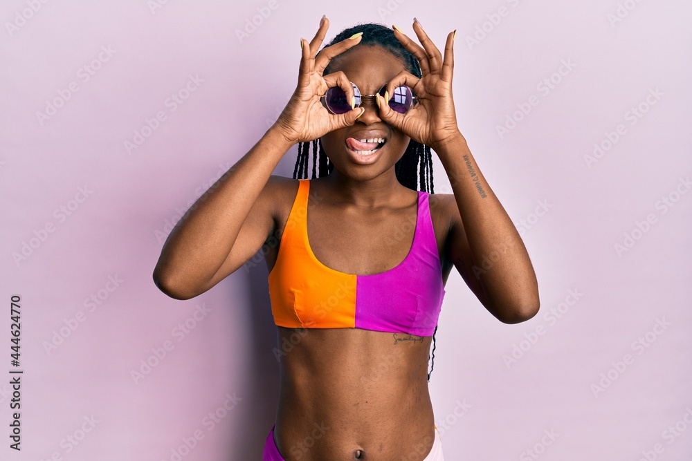 Young african american woman wearing bikini and sunglasses doing ok gesture like binoculars sticking tongue out, eyes looking through fingers. crazy expression. Young african american woman wearing bikini and sunglasses doing ok gesture like binoculars sticking tongue out, eyes looking through fingers. crazy expression.