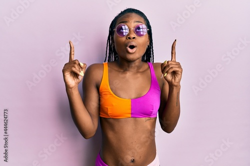 Young african american woman wearing bikini and sunglasses amazed and surprised looking up and pointing with fingers and raised arms.