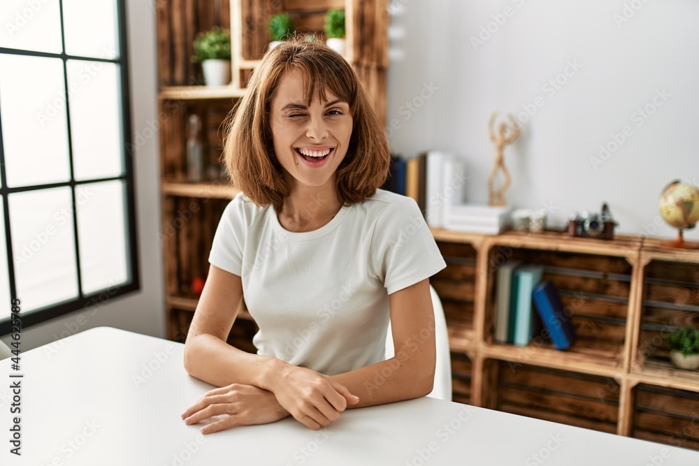 Young caucasian girl wearing casual clothes sitting on the table at home winking looking at the camera with sexy expression, cheerful and happy face. Young caucasian girl wearing casual clothes sitting on the table at home winking looking at the camera with sexy expression, cheerful and happy face.