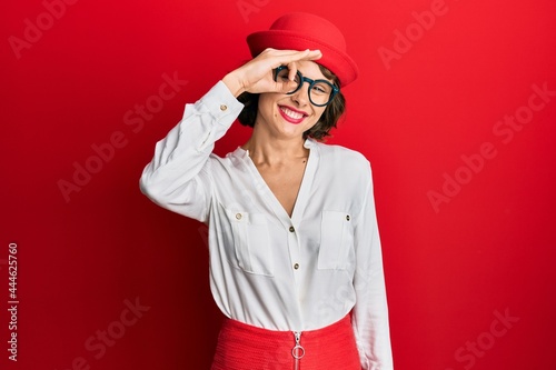 Young brunette woman wearing stewardess style and glasses smiling happy doing ok sign with hand on eye looking through fingers