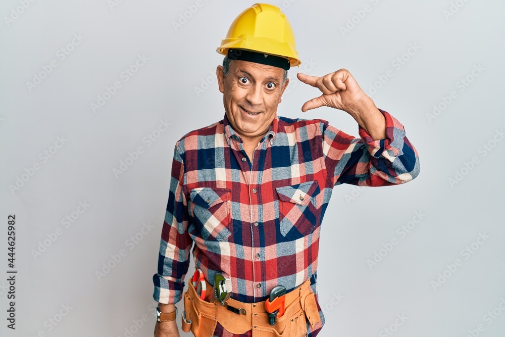 Senior hispanic man wearing handyman uniform smiling and confident gesturing with hand doing small size sign with fingers looking and the camera. measure concept.