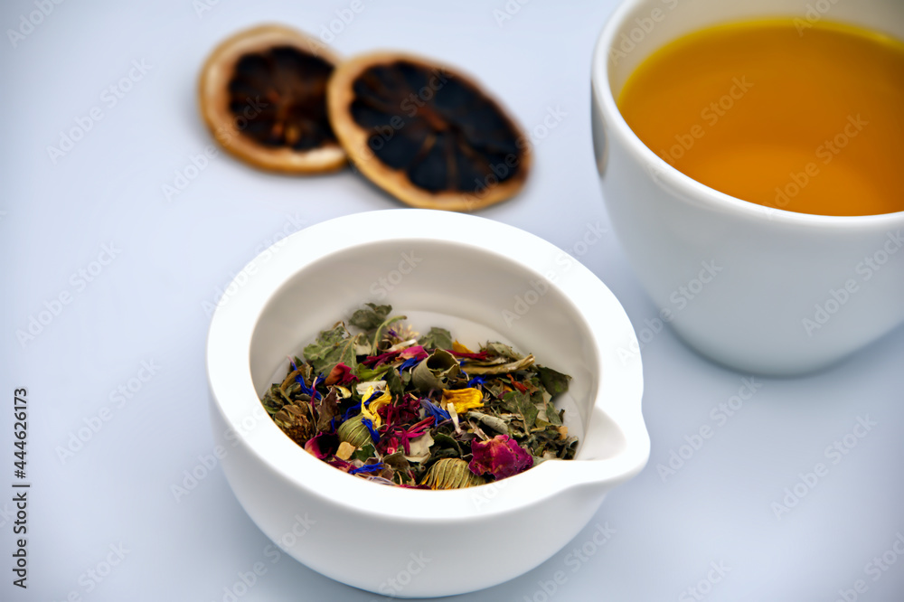 Dried herbs and flower petals in a white mortar, a cup of fresh tea and dried orange slices against a white background Dried herbs and flower petals in a white mortar, a cup of fresh tea and dried orange slices against a white background