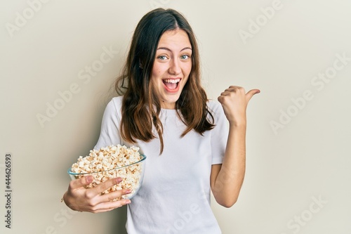 Young brunette woman eating popcorn pointing thumb up to the side smiling happy with open mouth