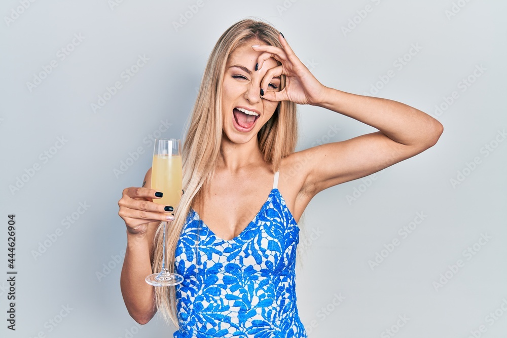 Young caucasian woman drinking a glass of sparkling champagne smiling happy doing ok sign with hand on eye looking through fingers Young caucasian woman drinking a glass of sparkling champagne smiling happy doing ok sign with hand on eye looking through fingers
