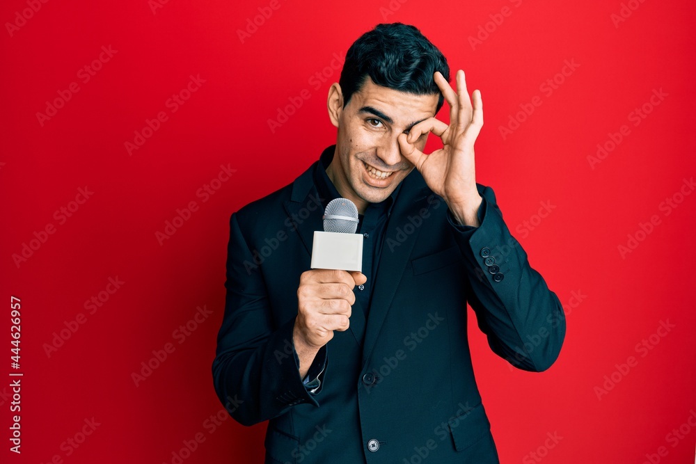 Handsome hispanic man holding reporter microphone smiling happy doing ok sign with hand on eye looking through fingers