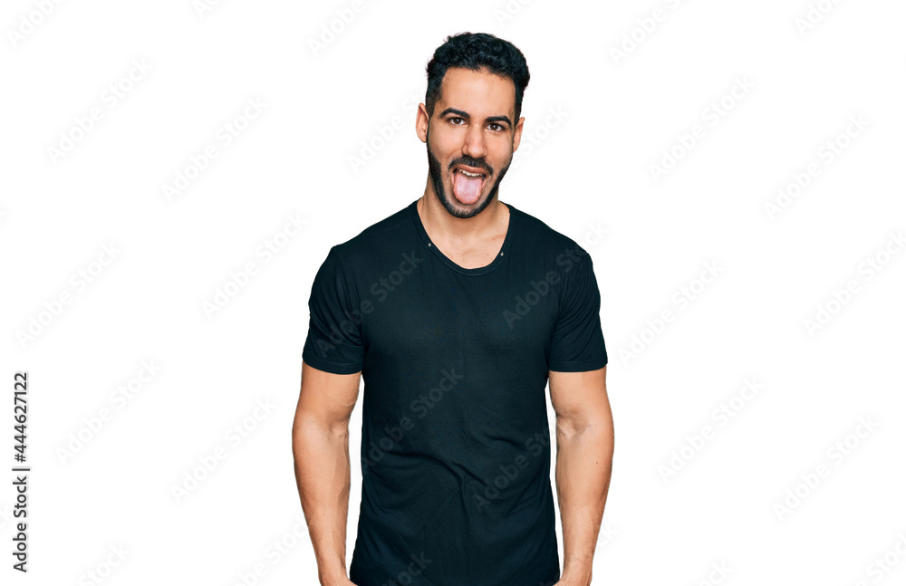 Hispanic man with beard wearing casual black t shirt sticking tongue out happy with funny expression. emotion concept. Hispanic man with beard wearing casual black t shirt sticking tongue out happy with funny expression. emotion concept.
