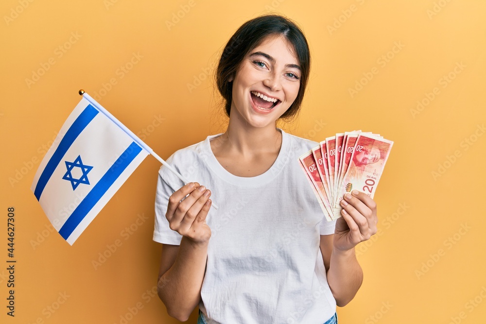 Young caucasian woman holding israel flag and shekels banknotes smiling and laughing hard out loud because funny crazy joke. Young caucasian woman holding israel flag and shekels banknotes smiling and laughing hard out loud because funny crazy joke.
