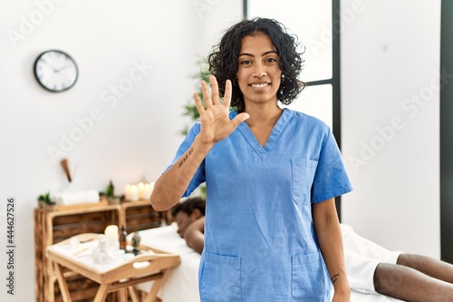 Young therapist woman at wellness spa center showing and pointing up with fingers number five while smiling confident and happy.