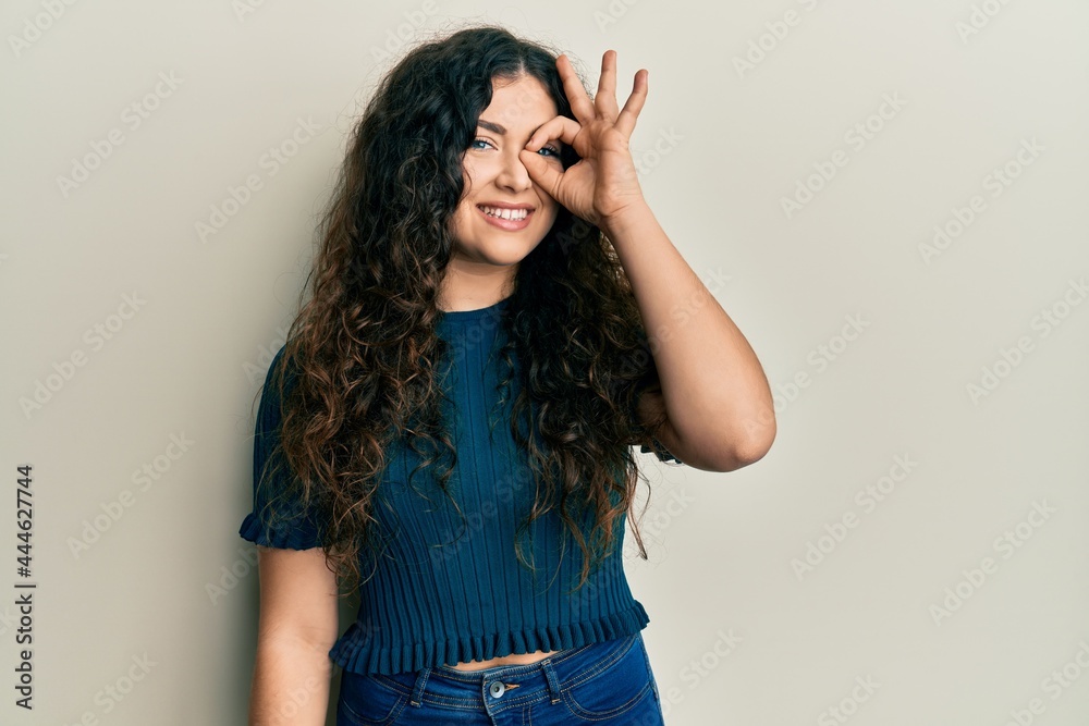 Young brunette woman with curly hair wearing casual clothes doing ok gesture with hand smiling, eye looking through fingers with happy face. Young brunette woman with curly hair wearing casual clothes doing ok gesture with hand smiling, eye looking through fingers with happy face.