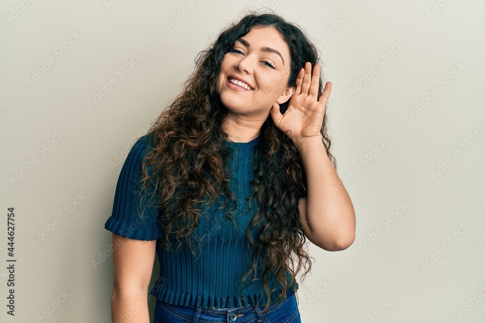 Young brunette woman with curly hair wearing casual clothes smiling with hand over ear listening an hearing to rumor or gossip. deafness concept. Young brunette woman with curly hair wearing casual clothes smiling with hand over ear listening an hearing to rumor or gossip. deafness concept.