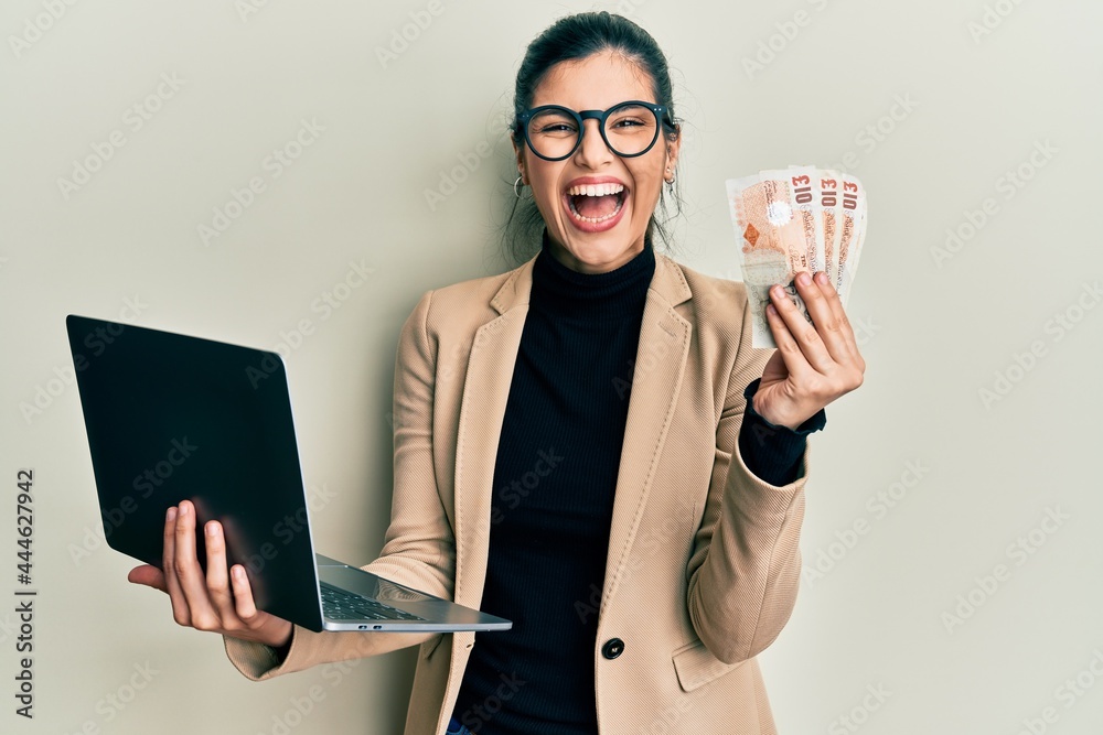 Young hispanic woman wearing business style holding laptop and united kingdom pounds celebrating crazy and amazed for success with open eyes screaming excited. Young hispanic woman wearing business style holding laptop and united kingdom pounds celebrating crazy and amazed for success with open eyes screaming excited.