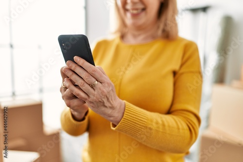 Middle age blonde woman smiling happy using smartphone at new home.