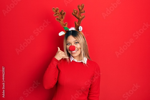 Beautiful hispanic woman wearing deer christmas hat and red nose smiling doing phone gesture with hand and fingers like talking on the telephone. communicating concepts.