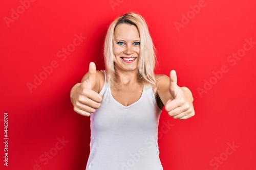 Beautiful caucasian blonde woman wearing casual white t shirt approving doing positive gesture with hand, thumbs up smiling and happy for success. winner gesture.