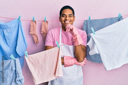 Young handsome hispanic man wearing cleaner apron holding clothes on clothesline looking confident at the camera with smile with crossed arms and hand raised on chin. thinking positive.