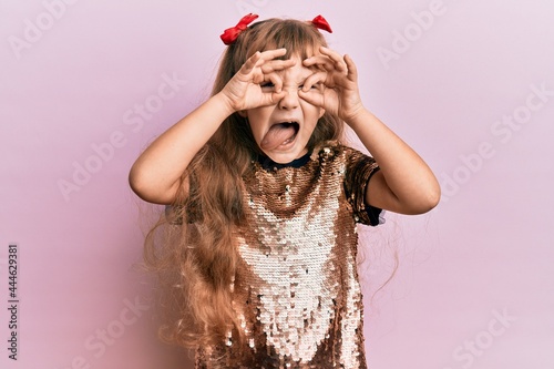 Little caucasian girl kid wearing festive sequins dress doing ok gesture like binoculars sticking tongue out, eyes looking through fingers. crazy expression.