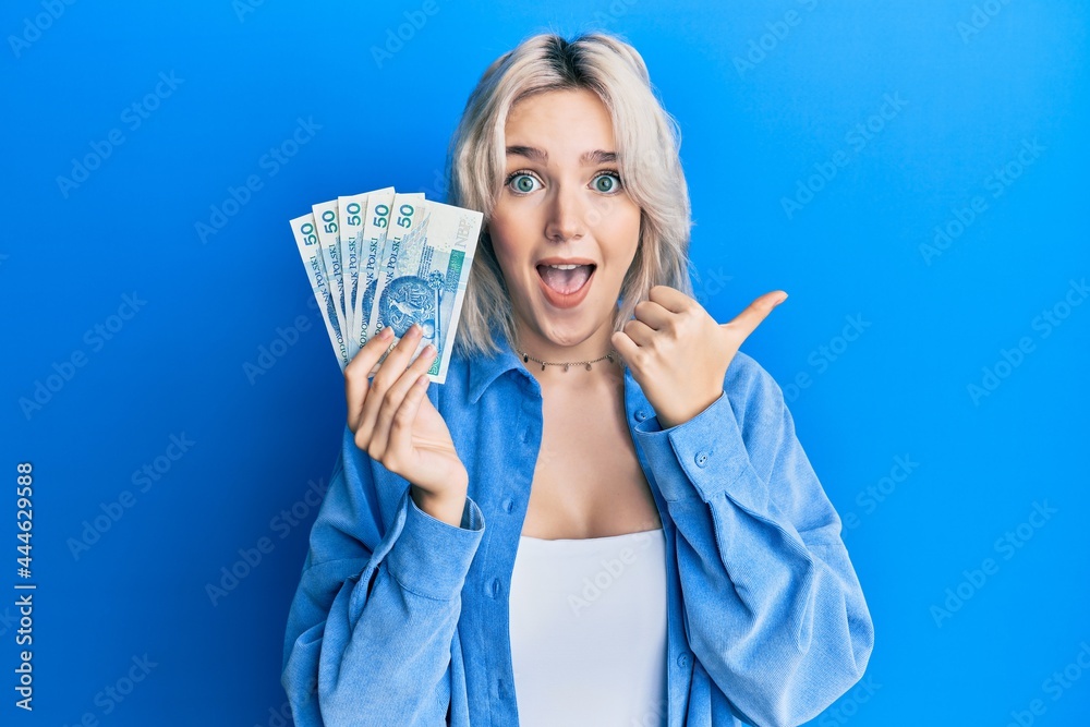 Young blonde girl holding 50 polish zloty banknotes pointing thumb up to the side smiling happy with open mouth Young blonde girl holding 50 polish zloty banknotes pointing thumb up to the side smiling happy with open mouth