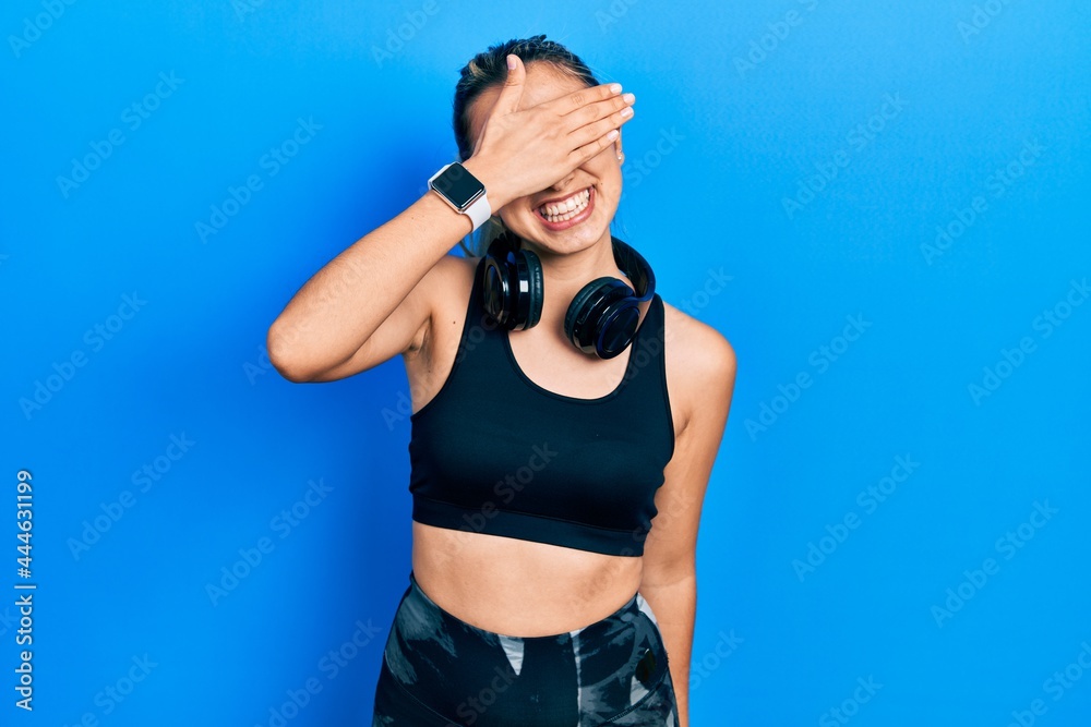 Beautiful hispanic woman wearing gym clothes and using headphones smiling and laughing with hand on face covering eyes for surprise. blind concept.