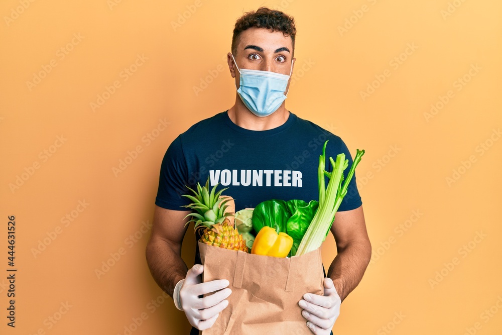 Hispanic young man wearing medical mask holding paper bag with groceries celebrating crazy and amazed for success with open eyes screaming excited. Hispanic young man wearing medical mask holding paper bag with groceries celebrating crazy and amazed for success with open eyes screaming excited.