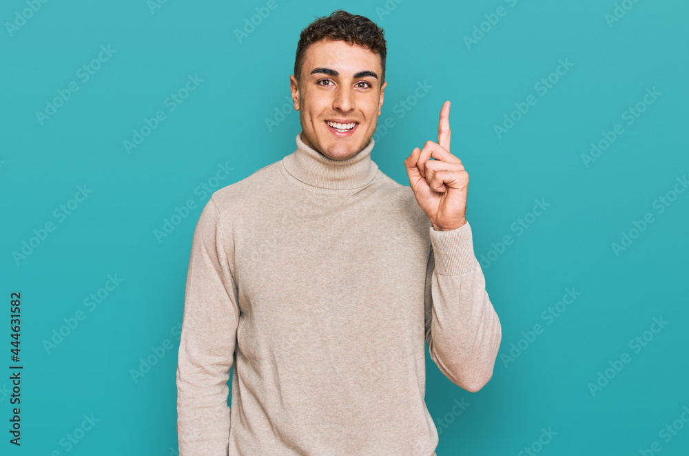 Fototapeta premium Hispanic young man wearing casual turtleneck sweater pointing finger up with successful idea. exited and happy. number one.