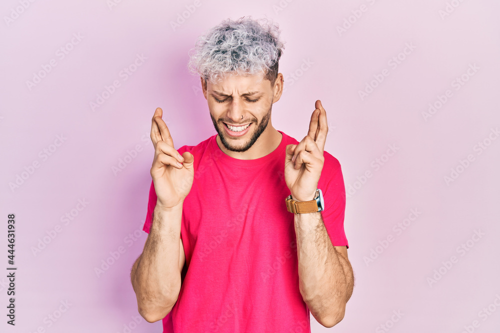 Young hispanic man with modern dyed hair wearing casual pink t shirt gesturing finger crossed smiling with hope and eyes closed. luck and superstitious concept. Young hispanic man with modern dyed hair wearing casual pink t shirt gesturing finger crossed smiling with hope and eyes closed. luck and superstitious concept.
