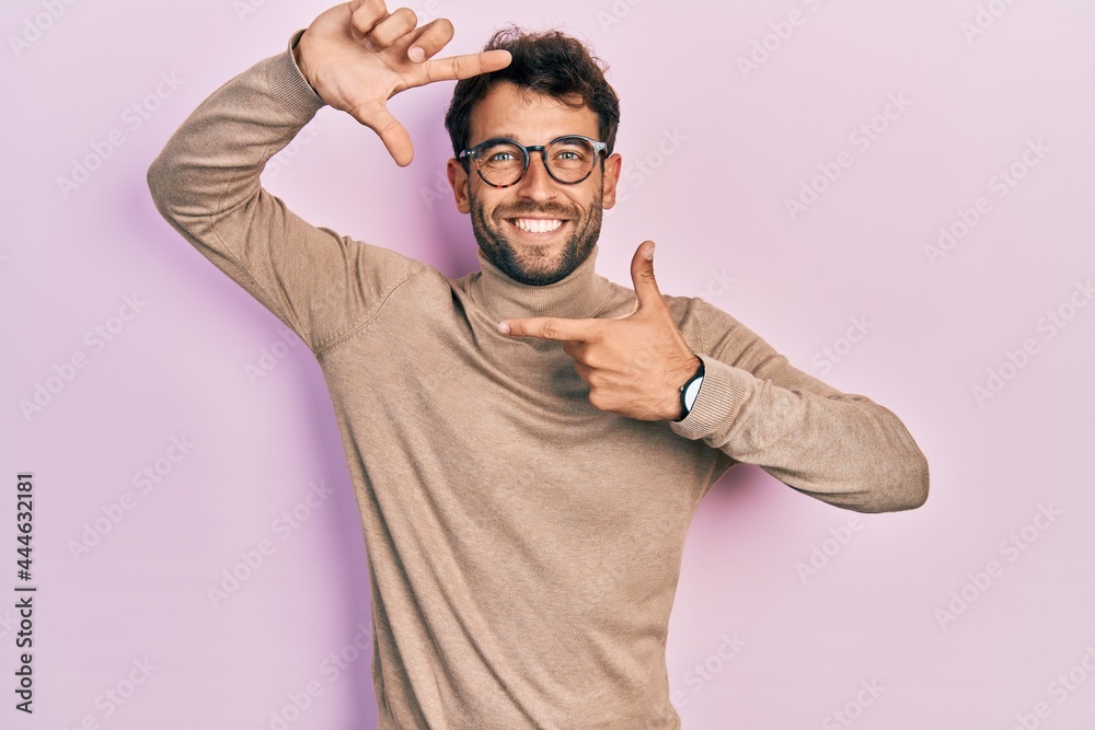 Handsome man with beard wearing turtleneck sweater and glasses smiling making frame with hands and fingers with happy face. creativity and photography concept.