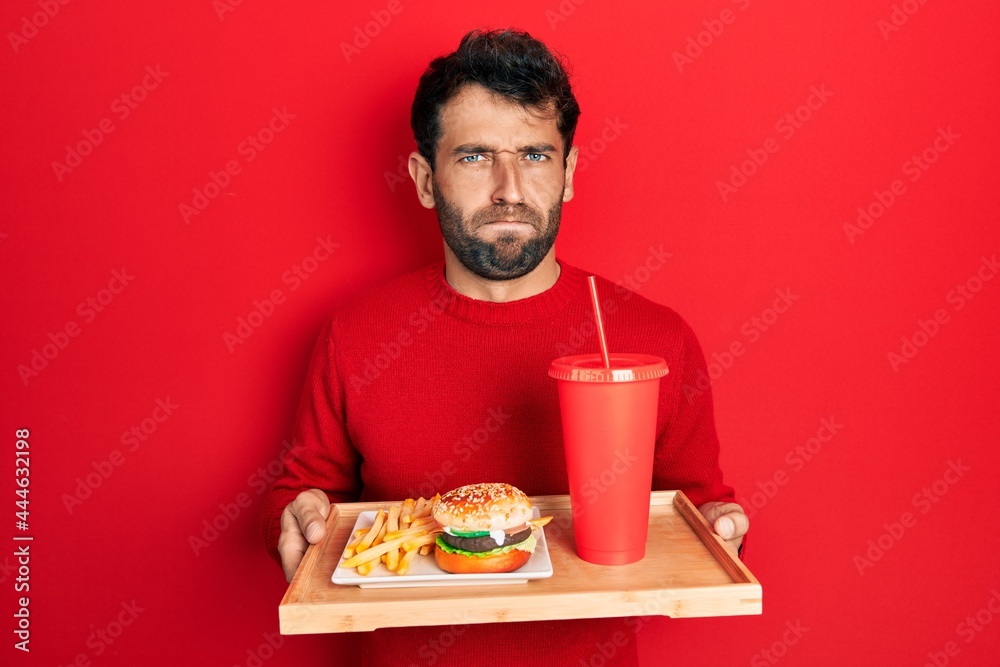 Handsome man with beard eating a tasty classic burger with fries and soda skeptic and nervous, frowning upset because of problem. negative person. Handsome man with beard eating a tasty classic burger with fries and soda skeptic and nervous, frowning upset because of problem. negative person.