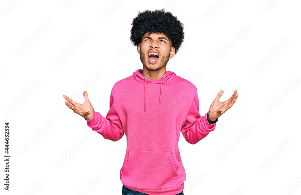 Young african american man with afro hair wearing casual pink sweatshirt crazy and mad shouting and yelling with aggressive expression and arms raised. frustration concept. Young african american man with afro hair wearing casual pink sweatshirt crazy and mad shouting and yelling with aggressive expression and arms raised. frustration concept.