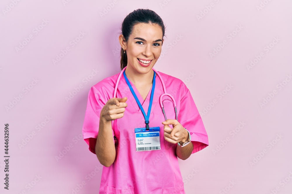 Young brunette woman wearing doctor uniform and stethoscope pointing fingers to camera with happy and funny face. good energy and vibes. Young brunette woman wearing doctor uniform and stethoscope pointing fingers to camera with happy and funny face. good energy and vibes.