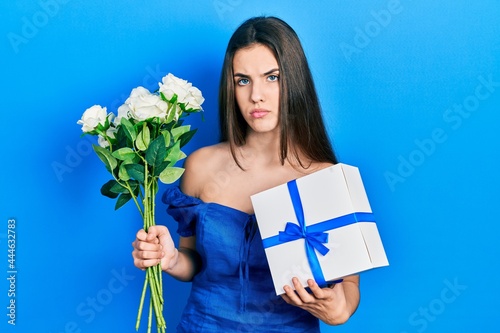 Young brunette teenager holding anniversary present and bouquet of flowers relaxed with serious expression on face. simple and natural looking at the camera.