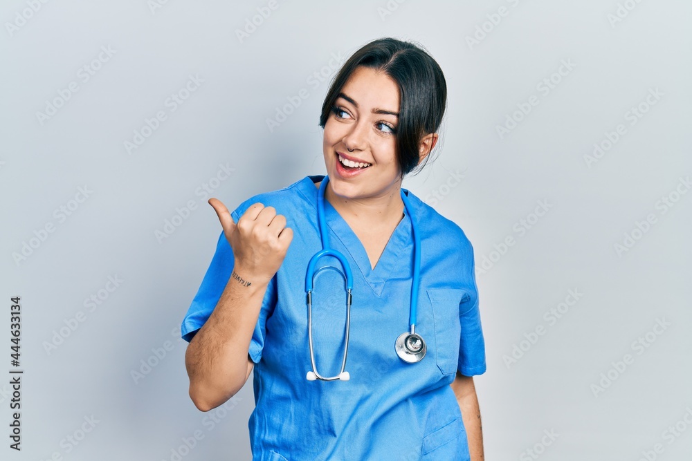 Beautiful hispanic woman wearing doctor uniform and stethoscope pointing thumb up to the side smiling happy with open mouth Beautiful hispanic woman wearing doctor uniform and stethoscope pointing thumb up to the side smiling happy with open mouth