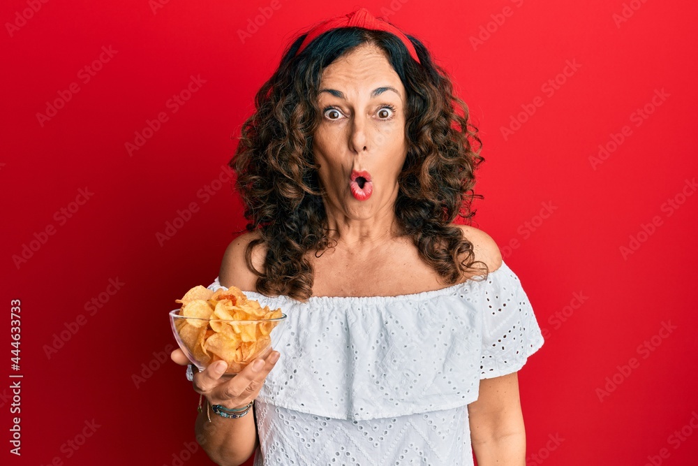 Middle age hispanic woman holding potato chip scared and amazed with open mouth for surprise, disbelief face Middle age hispanic woman holding potato chip scared and amazed with open mouth for surprise, disbelief face