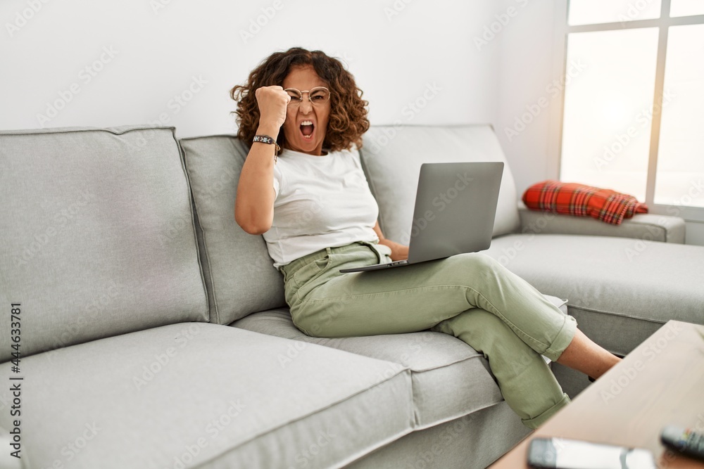 Middle age hispanic woman working using computer laptop at home annoyed and frustrated shouting with anger, yelling crazy with anger and hand raised Middle age hispanic woman working using computer laptop at home annoyed and frustrated shouting with anger, yelling crazy with anger and hand raised