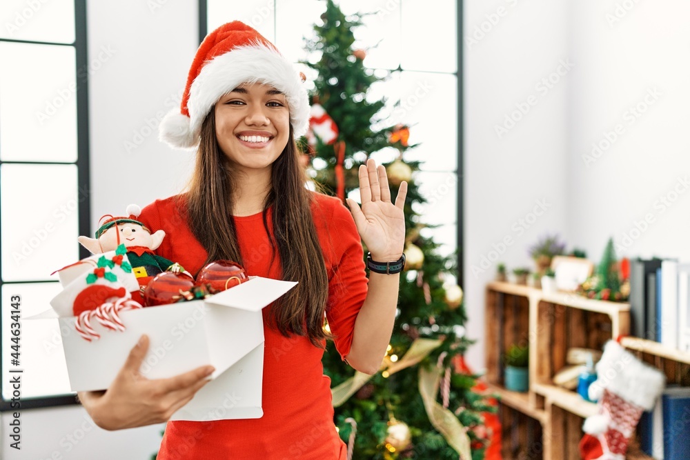 Young brunette woman standing by christmas tree holding decoration waiving saying hello happy and smiling, friendly welcome gesture Young brunette woman standing by christmas tree holding decoration waiving saying hello happy and smiling, friendly welcome gesture