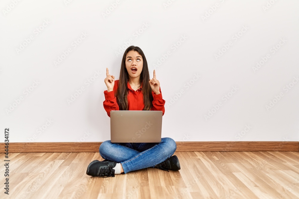 Young brunette woman sitting on the floor at empty room with laptop amazed and surprised looking up and pointing with fingers and raised arms. Young brunette woman sitting on the floor at empty room with laptop amazed and surprised looking up and pointing with fingers and raised arms.