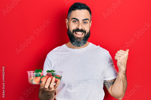 Young hispanic man holding strawberries pointing thumb up to the side smiling happy with open mouth