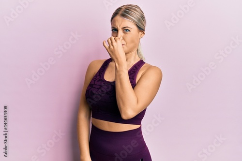 Beautiful blonde woman wearing sportswear over pink background smelling something stinky and disgusting  intolerable smell  holding breath with fingers on nose. bad smell