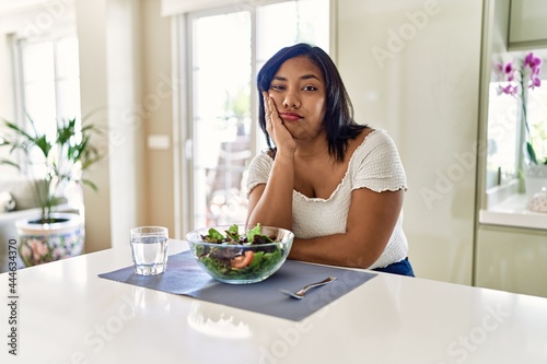 Young hispanic woman eating healthy salad at home thinking looking tired and bored with depression problems with crossed arms.