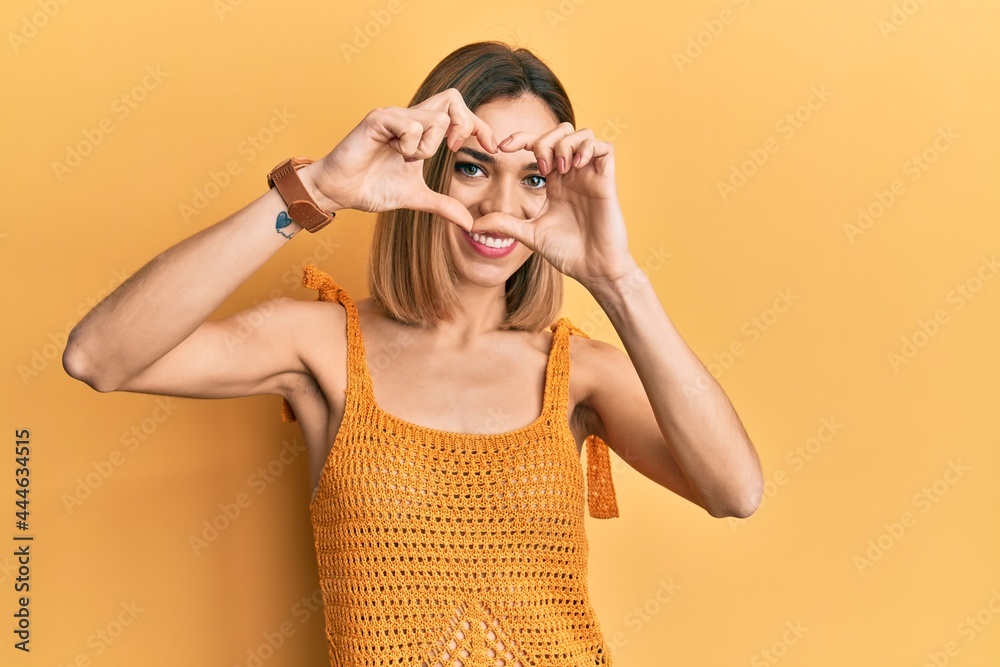 Young caucasian blonde woman wearing casual yellow t shirt doing heart shape with hand and fingers smiling looking through sign Young caucasian blonde woman wearing casual yellow t shirt doing heart shape with hand and fingers smiling looking through sign