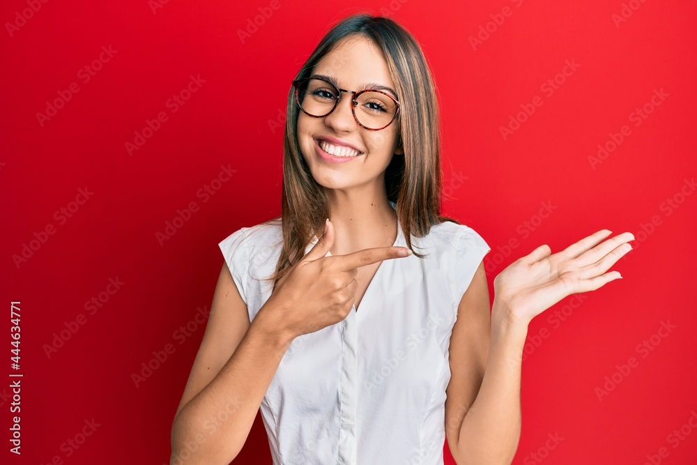 Young brunette woman wearing casual clothes and glasses amazed and smiling to the camera while presenting with hand and pointing with finger. Young brunette woman wearing casual clothes and glasses amazed and smiling to the camera while presenting with hand and pointing with finger.