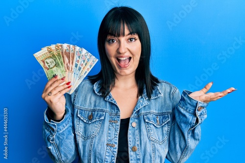 Young hispanic woman holding peruvian sol banknotes celebrating achievement with happy smile and winner expression with raised hand