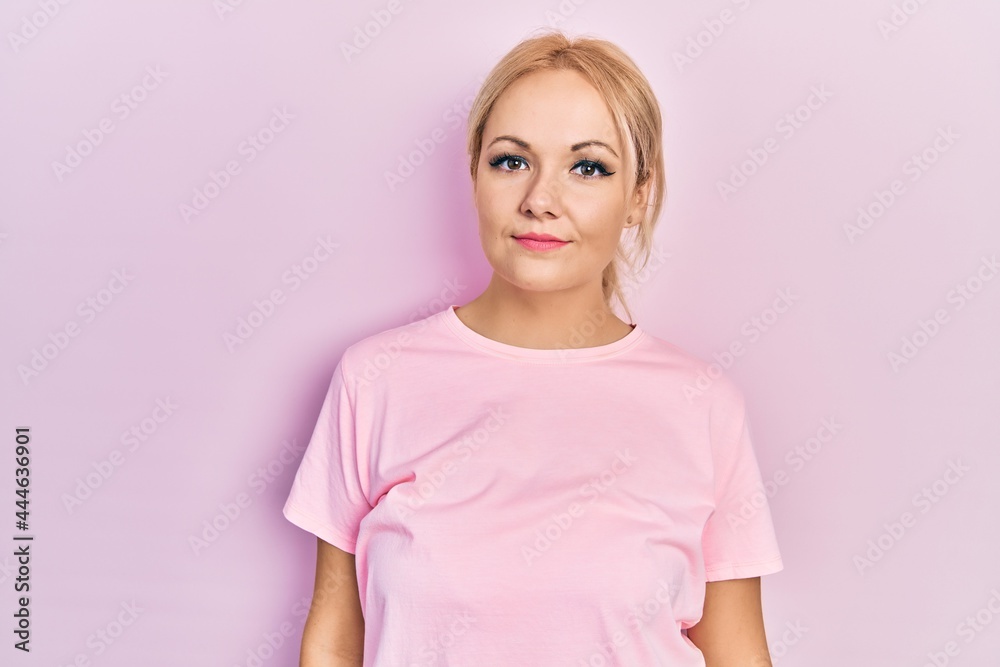 Young blonde woman wearing casual pink t shirt relaxed with serious expression on face. simple and natural looking at the camera. Young blonde woman wearing casual pink t shirt relaxed with serious expression on face. simple and natural looking at the camera.