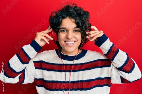 Young hispanic woman with curly hair listening to music using headphones smiling with a happy and cool smile on face. showing teeth.