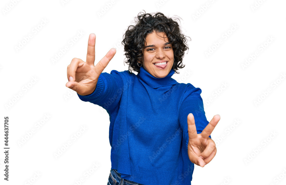 Young hispanic woman with curly hair wearing turtleneck sweater smiling with tongue out showing fingers of both hands doing victory sign. number two. Young hispanic woman with curly hair wearing turtleneck sweater smiling with tongue out showing fingers of both hands doing victory sign. number two.